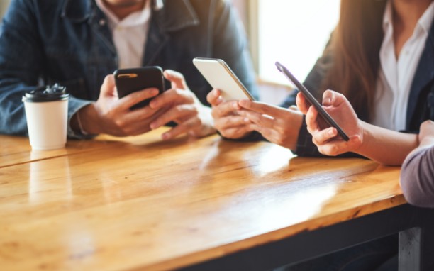 Três pessoas sentadas numa mesa de madeira entunato seguram e observam a tela dos seus celulares.