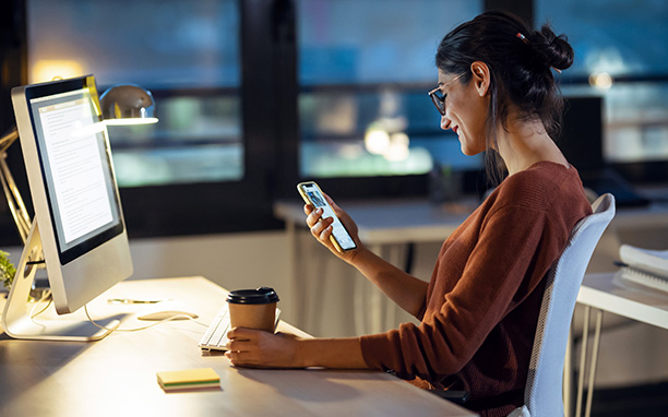 Mulher sentada encostada em uma cadeira segurando um copo de café em uma mão enquanto segura um celular com a outra e  observa com um sorriso.