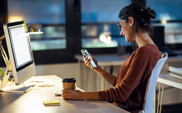 Mulher sentada encostada em uma cadeira segurando um copo de café em uma mão enquanto segura um celular com a outra e observa com um sorriso.
