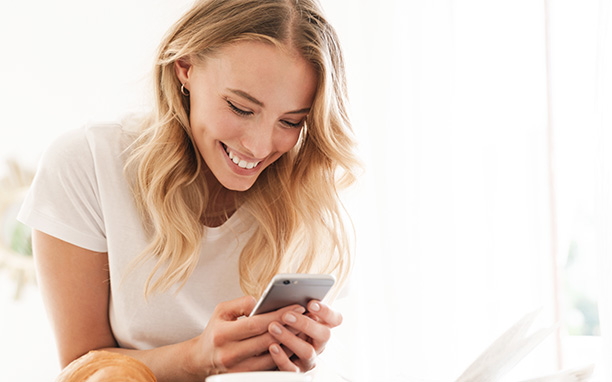 Mulher branca loira sorri ao observar a tela de um celular.