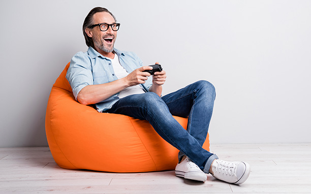 Homem de meia idade, sentado em um pufe, com um joystick sem fio e com semblante de euforia.