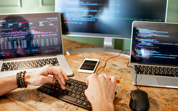 Em uma mesa, homem digitando em um teclado externo, dois notebooks aberto e um smartphone no centro.